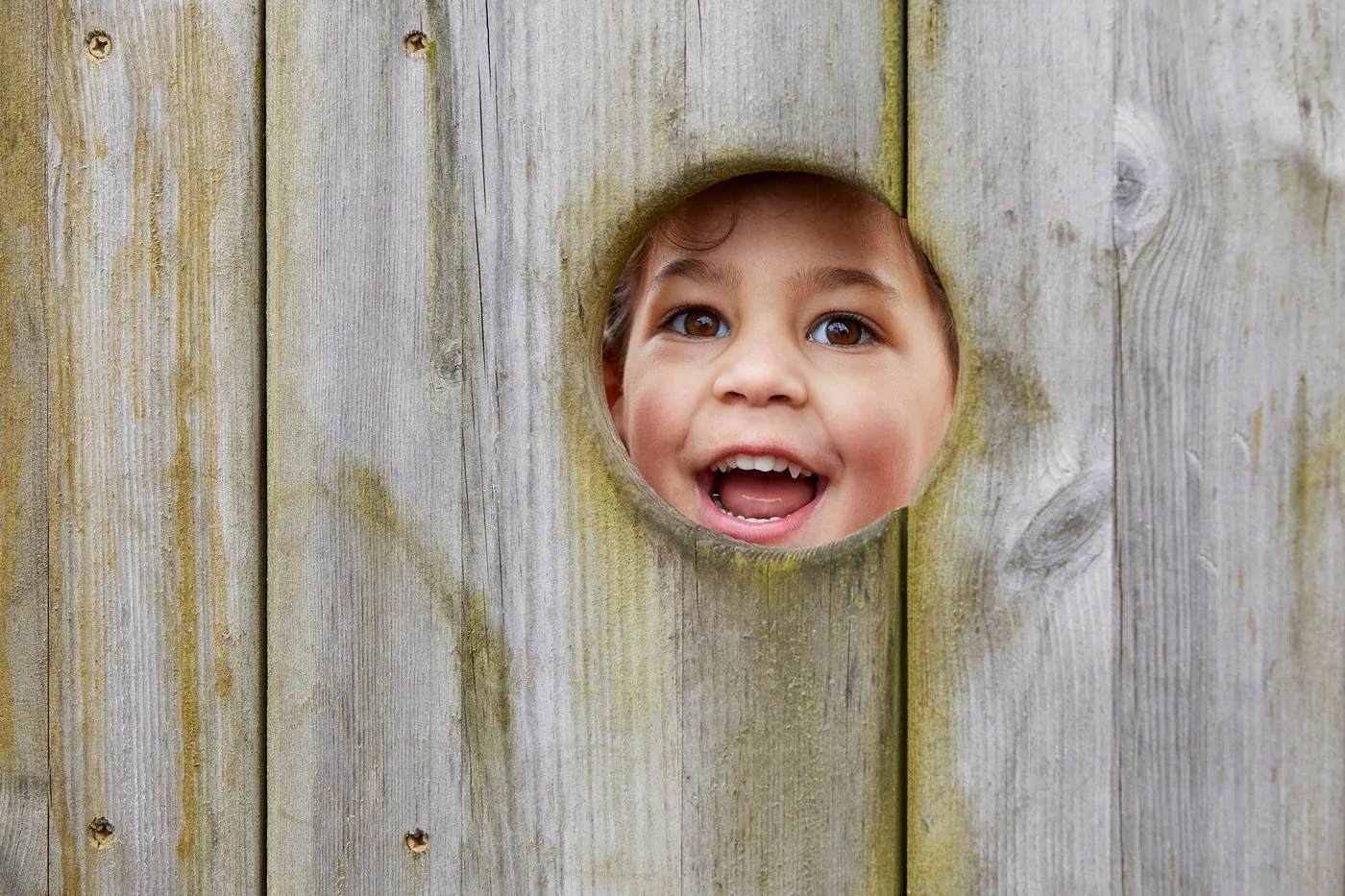 A smiling child looking through a round hole in a wooden fence